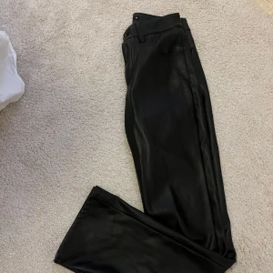 Svarta raka byxor i skinnimitation - Snygga svarta byxor i skinnimitation med raka ben och klassisk femficksmodell. Perfekta för dig som vill ha en edgy look och gillar stilrena detaljer. Byxorna har hällor för bälte och stängs med dragkedja och knapp.