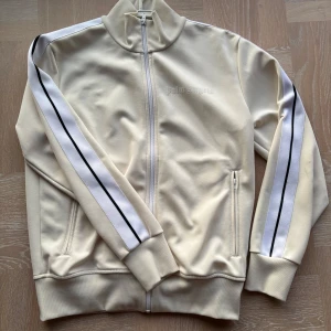 Palm Angels vit track jacket - Palm Angels track jacket i offwhite med svarta och vita stripes längs ärmarna. Säljer min kofta i väldigt unik och sällsynt färg. Endast en flaw som är synlig som man ser i sista bilden annars i nästan nyskick. Tags kan eventuellt finnas om jag hittar dem, självklart Äkta. Dm för frågor eller prisförslag