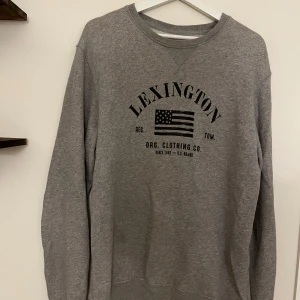 Grå sweatshirt från Lexington - Grå sweatshirt från Lexington med svart tryck och amerikansk flagga på bröstet. Klassisk rund halsringning och långa ärmar. Perfekt för en avslappnad och stilren look.