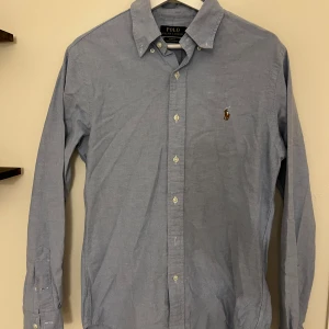 Blå skjorta från Polo Ralph Lauren - Klassisk blå skjorta från Polo Ralph Lauren med broderad logga på bröstet. Skjortan har lång ärm, knappar hela vägen och button-down krage. Perfekt för en clean och stilren look.