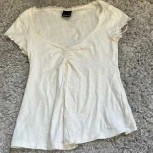 Vit blus med spetsdetaljer - Söt vit blus från Monki med v-ringning och fina spetsdetaljer längs halsringningen. Toppen har korta ärmar och en lätt rynkning framtill som ger en feminin touch. Perfekt för dig som gillar en romantisk och enkel stil.