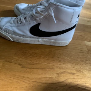 Nike Blazer Mid vita sneakers  - Snygga vita Nike Blazer Mid sneakers med svart swoosh och klassisk retrostil. Skorna har högt skaft, snörning och en robust sula. Perfekta för dig som gillar streetwear och vill ha en tidlös look.