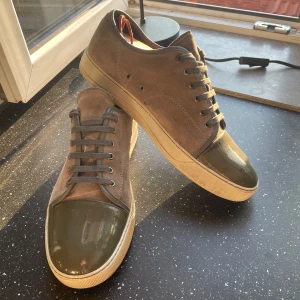 Lanvin sneakers i mocka och lack - Snygga sneakers från Lanvin i brun mocka med olivgrön lackad tå och matchande snörning. Klassisk siluett med rund tå och beige sula. Perfekta för dig som vill ha en stilren men unik look med exklusiv känsla.