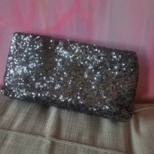 Silverglittrig clutch från Victoria's Secret - Clutch från Victoria's Secret täckt i glittrande silverpaljetter. Rektangulär form med magnetknäppning och svart innerfoder. Logga i metall framtill för extra bling. Så så fin till en utekväll!! Använd en gång så i nyskick! Går inte att köpa längre 🩷💖