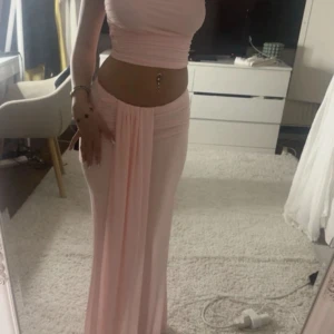 Ljusrosa sett - Supersnygg ljusrosa långkjol med bandeau-topp och figurnära passform. Settet har en draperad detalj framtill i midjan och är axelbandslös, vilket ger en clean och modern look. Perfekt för dig som vill sticka ut med en feminin och trendig stil.