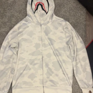 Vit camo hoodie från Bape - Säljer en vit hoodie från Bape med grått camo-mönster och ikonisk hajdesign på huvan. Tröjan har dragkedja hela vägen, känguruficka och ribbade muddar. Perfekt statement-plagg för dig som gillar streetwear.
