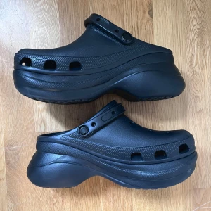 Svarta Crocs BAE med platå, nya - Svarta Crocs, modell BAE som är en variant av den klassiska modellen fast med platåsula. Nyinköpta och har endast använts en gång och säljer för att de är för stora. Strl är W9, vilket motsvarar EU 40. Gör ett klipp och bli coolast i stan!