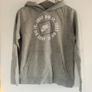 Grå Nike hoodie med tryck - Snygg grå hoodie från Nike med klassiskt 'Just Do It'-tryck i vitt på bröstet. Tröjan har huva, magficka och långa ärmar. Perfekt för chill dagar eller när du vill ha en sportig vibe. Mjuk bomullsblandning som känns skön mot huden.