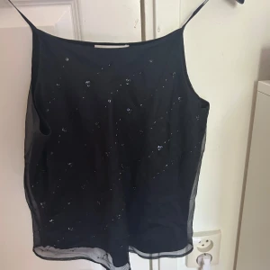 Marin blå glittrig linnetopp med paljetter - Säljer en svart linnetopp med tunna axelband och glittriga paljettdetaljer framtill. Toppen är lätt och luftig med ett transparent lager ovanpå, perfekt för dig som gillar en stilren men ändå festlig look.