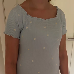 Ljusblå blommig kortklänning - Supersöt ljusblå ribbad kortklänning med små broderade blommor i vitt och gult. Klänningen har korta ärmar, vågig kant vid halsringning och ärmslut samt en figurnära passform. Perfekt för sommarens alla tillfällen!