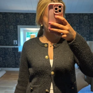 Mörkgrå cardigan jacka med guldknappar från Zara - Mysig mörkgrå cardigan jacka med snygga guldknappar framtill och två små fickor. Jackan är stickad och har en klassisk rund halsringning samt lång ärm. Perfekt att slänga över en topp för en chill men ändå uppklädd look.