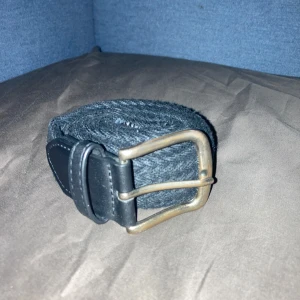 Svart flätat bälte med metallspänne - Snyggt svart flätat bälte med robust metallspänne i matt mässingsfärg. Bältet har detaljer i svart skinn vid ändarna och passar perfekt till jeans eller chinos. Klassisk design som funkar till många olika stilar.