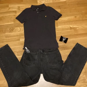 Svart pikétröja från Lyle & Scott - Klassisk svart pikétröja från Lyle & Scott Junior med gul broderad logga på bröstet. Tröjan har krage, knappar framtill och korta ärmar. Perfekt för en clean och stilren look. Tillverkad i mjuk bomull som känns skön mot huden. Ändast tröjan medföljer vid köp