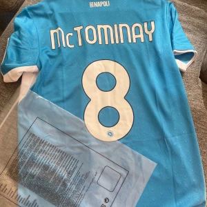 Napoli McTominay matchtröja blå - Snygg Napoli fotbollströja med McTominay och nummer 8 på ryggen. Tröjan är blå med vita detaljer, har klassisk krage och korta ärmar. Märkt med EA7 och MSC-logga framtill. Tillverkad i lätt och ventilerande material, perfekt för match eller träning.
