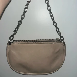 Beige axelväska med kedja - Begie axelväska i skinnimitation med grov silverfärgad kedja som axelrem. Väskan har rundad form och stängs med dragkedja upptill. Insidan är svartfodrad.