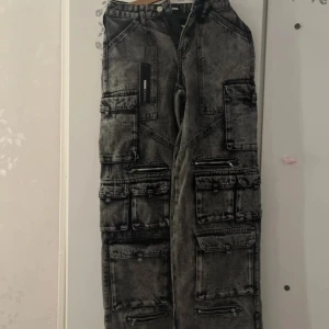 Gråsvarta cargo jeans från Fashion Nova - Riktigt feta gråsvarta cargo jeans från Fashion Nova med tvättad look. Massor av fickor och dragkedjor ger en cool streetstyle-vibe. Vida ben och loose fit, tillverkade i jeansmaterial. Perfekta för dig som gillar utility och edgy stil.