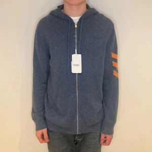 Blå hoodiejacka från Zadig & Voltaire - Blå hoodiejacka från Zadig & Voltaire med dragkedja och huva. Jackan har tre orangea ränder på ena ärmen och är tillverkad i mjukt material. Perfekt för dig som gillar en chill och stilren look.
