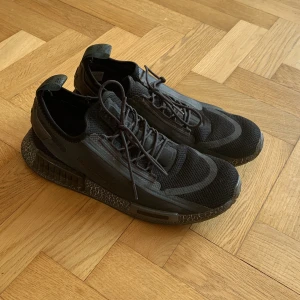 Svarta Adidas NMD sneakers - Säljer ett par svarta Adidas NMD sneakers med coolt mönstrad sula och mesh-överdel. Skorna har snörning, diskreta Adidas-loggor och detaljer i syntet. Perfekta för dig som gillar streetwear och vill ha en unik look.