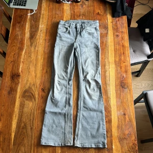 Grå bootcut jeans med dragkedja strl 146 - Grå jeans med bootcut passform och klassisk femficksdesign. Jeansen har dragkedja och knapp framtill samt bälteshällor. Mjuka och stretchiga. 