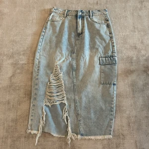 Lång jeanskjol med slitningar - lång jeanskjol i ljusblå denim med stor sliten detalj framtill och fransad nederkant. Kjolen har cargoficka på sidan och klassiska jeansfickor. I gott skick och änvänd 2-3 gånger. Storlek M. Säljs för 45kr 