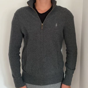 Mörkgrå zip från Polo Ralph Lauren - Mörkgrå zip från Polo Ralph Lauren med hel dragkedja och ståkrage. Tröjan är stickad i mjuk ull och har diskret logga broderad på bröstet. Perfekt för lager-på-lager och chill stil. Klassisk och enkel design som funkar till det mesta.