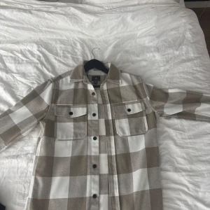 Rutig overshirt från H&M i beige och vitt - Overshirt från H&M i stor rutig design med beige och vita toner. Klassisk skjortkrage, två bröstfickor med knappar och svarta knappar framtill. Perfekt lager-på-lager-plagg för en chill och trendig look.