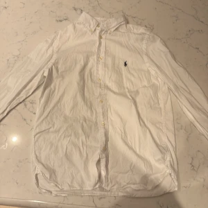 Vit skjorta från Polo Ralph Lauren - Klassisk vit skjorta från Polo Ralph Lauren i slim fit-modell. Skjortan har långa ärmar, knappar framtill och den ikoniska broderade loggan på bröstet. Tillverkad i mjuk bomull och passar perfekt till en clean och stilren look.