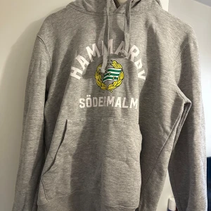 Grå Hammarby hoodie med tryck - Grå hoodie från Hammarby med stort tryck på bröstet där det står 'HAMMARBY SÖDERMALM' och klubbens emblem i grönt, gult och vitt. Tröjan har huva med snörning och en stor magficka. Perfekt för dig som vill visa ditt stöd för laget.