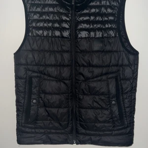 Svart dunväst från BOSS - Snygg svart dunväst från BOSS med quiltad design och dragkedja framtill. Västen har två fickor med knappstängning och diskret BOSS-logga på bröstet. Insidan är fodrad i beige och orange detaljer. Perfekt för lager-på-lager-stil.