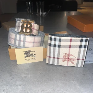 Burberry rutig bälte med guldspänne - Säljer ett snyggt Burberry-bälte i klassiskt beige, vitt och grått rutigt mönster. Bältet har ett stort, guldigt B-spänne framtill och är gjort i skinn. Perfekt accessoar för att lyfta din outfit med en lyxig touch.