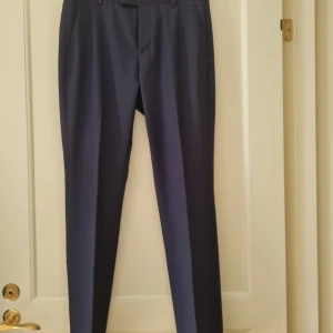 Mörkblå kostymbyxor från H&M strl 46 - Snygga mörkblå kostymbyxor från H&M i slim fit. Perfekta för en skolavslutning. Byxorna har en klassisk design med knappar bak och dragkedja fram. Storlek 46. Kan hämtas i Vallentuna.