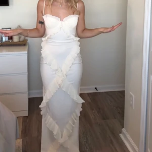 Vit långklänning med volangdetaljer OHPOLLY - Supersnygg vit långklänning med smala axelband och markerad byst. Klänningen har volangdetaljer i ett X-mönster framtill som ger en unik look. Figurnära passform och romantisk vibe, perfekt för dig som vill sticka ut.