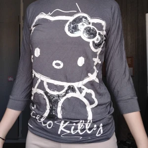 Grå Hello Kitty tröja från H&M - Grå långärmad tröja från H&M med stort Hello Kitty-tryck framtill och paljettdetaljer på rosetten och klänningen. Tröjan har trekvartsärm och är gjord i mjuk bomull, perfekt för dig som gillar statement-plagg med lite extra bling.