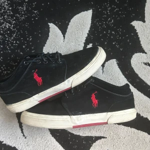 Polo Ralph Lauren skor - Svarta sneakers från Polo Ralph Lauren med röd broderad logga på sidan och vit sula med röd undersida. Skorna har snörning och är tillverkade i mocka och textil. Klassisk och stilren modell med rund tå och diskreta detaljer.