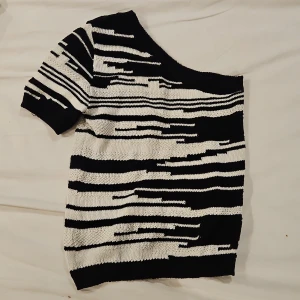 Randig offshoulder stickad tröja XS - Snygg stickad tröja från Lindex i svart och vitt med coolt randigt mönster. Tröjan har offshoulder-design och korta ärmar, perfekt för dig som vill sticka ut lite extra. Gjord i återvunnen bomullsblandning för en skön känsla.