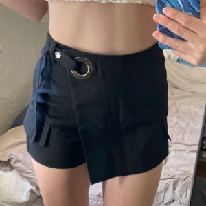 Svarta omlottshorts med ringdetalj - Snygga svarta shorts med omlott-design och stor silverfärgad ringdetalj i midjan. Shortsen har elastisk midja bak för extra komfort. Perfekta för en trendig och edgy look.