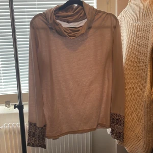 Beige långärmad tröja med mönstrade ärmar - Beige långärmad tröja med lös passform och snygg draperad krage. Ärmsluten har kontrasterande muddar med svartvitt mönster som ger en unik touch. Perfekt för dig som gillar stilrena men ändå personliga plagg.