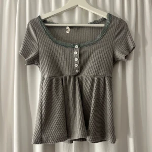 Grå ribbad blus från Romwe - Supersöt grå ribbad blus från Romwe med korta ärmar, knappar framtill och spetskant vid den vida urringningen. Modellen har en lätt utställd nederdel som ger en flowy känsla. Perfekt för dig som gillar en chill men ändå söt stil.