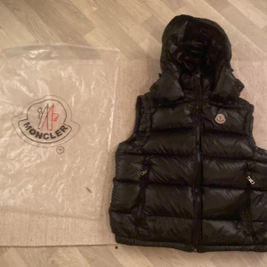 Svart dunväst från Moncler med huva - Säljer en svart dunväst från Moncler med avtagbar huva och klassisk Moncler-logga på bröstet. Västen har två dragkedjefickor, quiltad design och blanka ytor. Perfekt för lager-på-lager och riktigt snygg till streetwear-stilen.