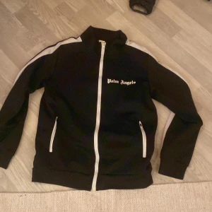 Palm Angels svart trackjacka med vita detaljer - Svart trackjacka från Palm Angels med vita ränder längs ärmarna och vit dragkedja. Jackan har hög krage, två fickor med dragkedja och logga på bröstet. Perfekt för en sportig och trendig streetstyle-look. Den e inte äkta 
