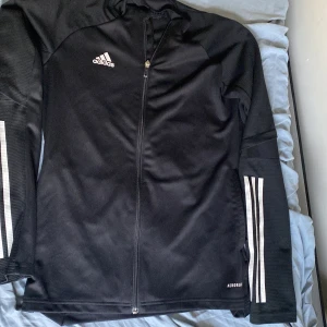 Svart Adidas träningsjacka S - Svart träningsjacka från Adidas med hel dragkedja och klassiska vita ränder på ärmarna. Tillverkad i lätt polyester med AEROREADY-teknologi som andas. Perfekt för träning eller chill. Adidas-logga på bröstet och smal passform.