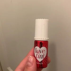 Benefit Love Tint läpp- och kindfärg - Röd flytande tint från Benefit i nyansen Love Tint. Kan användas både på läppar och kinder för en naturlig, fräsch look. Kommer i en liten röd flaska med vitt lock och dekorativa detaljer. Perfekt för att ge färg som håller hela dagen.