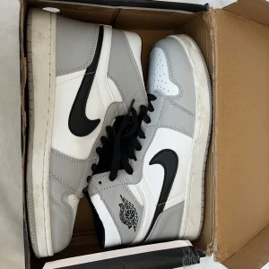 Nike Air Jordan 1 High vit/grå - Snygga Nike Air Jordan 1 High sneakers i vitt och ljusgrått med svarta detaljer och klassisk svart swoosh. Skorna har svart snörning, perforerad tå och ikonisk Air Jordan-logga på sidan. Tillverkade i skinn och syntet för en clean och sportig look.