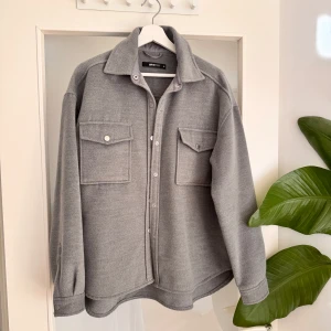 Grå overshirt från Gina Tricot - Snygg grå overshirt från Gina Tricot med klassisk krage, stora bröstfickor och knappar framtill. Oversized passform och mjukt material som ger en chill vibe. Använd sparsamt och är i bra skick utan defekter.