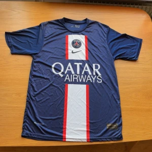 PSG Mbappé matchtröja Nike XL - Paris Saint-Germain fotbollströja med Mbappé och nummer 7 på ryggen. Tröjan är marinblå med vita och röda detaljer, PSG-emblem på bröstet och Qatar Airways som sponsor. Tillverkad i Dri-FIT-material som andas. Klassisk kortärmad modell.