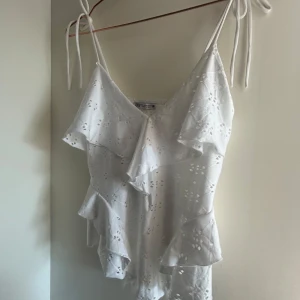 Vit volangblus från Zara - Superfin vit blus från Zara med tunna axelband som knyts på axlarna. Blusen har broderade hålmönster och flera volangdetaljer framtill och på sidorna. Perfekt för sommarens varma dagar och ger en romantisk vibe.