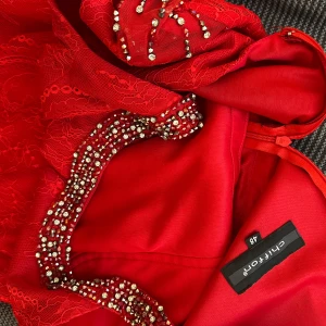 Röd klänning med spets och pärldetaljer från Chantana - Säljer en röd klänning från Chantana med vackra spetsdetaljer och pärlor längs axlar och midja. Klänningen har inslag av transparent mesh och figurnära passform. Perfekt för dig som vill sticka ut med elegans. Passar XXL 