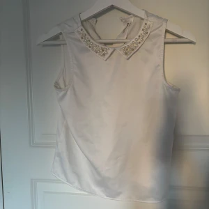 Vit ärmlös blus med pärldetaljer från H&M - Säljer en vit ärmlös blus från H&M med dekorativa pärlor och små guldfärgade detaljer på kragen. Blusen har en  krage och är av ett glansigt material💕