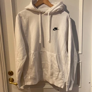 Vit hoodie från Nike - Säljer en klassisk vit hoodie från Nike med svart broderad logga på bröstet. Tröjan har huva med dragsko och en stor magficka framtill. Perfekt för en avslappnad och sportig stil.