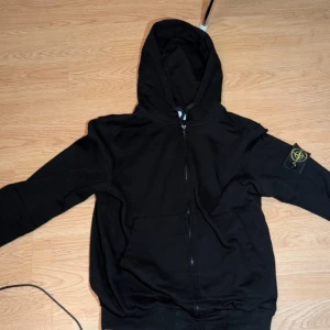 Svart hoodie från Stone Island - Säljer en svart hoodie från Stone Island med dragkedja och huva. Klassisk patch med logga på vänster ärm och två fickor framtill. Perfekt för en avslappnad och stilren look.